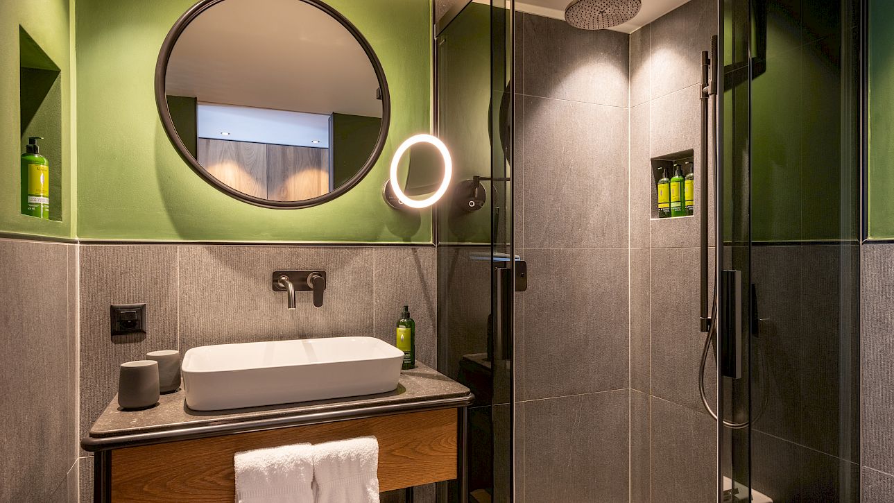 Modernes Badezimmer mit grün-grauen Wänden, rundem Spiegel, Waschtisch und verglaster Dusche.