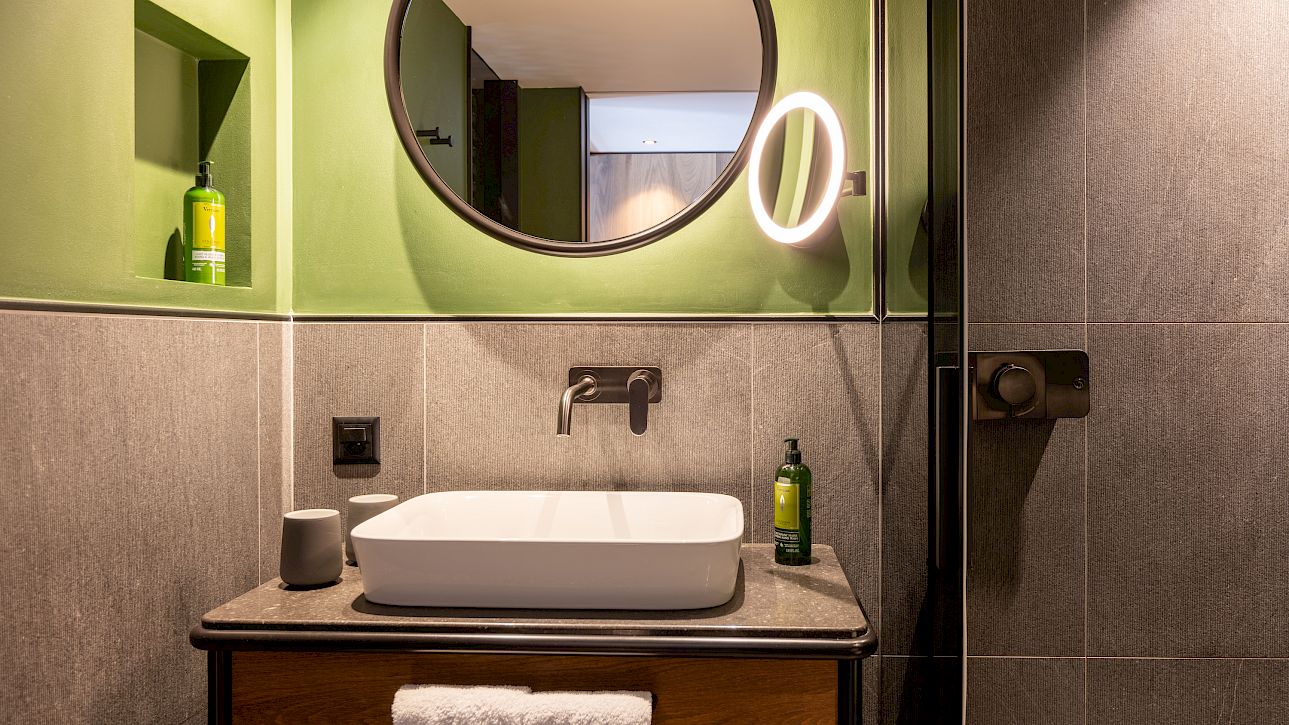 Modernes Badezimmer mit grünem Spiegel, beleuchtetem Wandspiegel und grauen Fliesen. Seifenspender vorhanden.