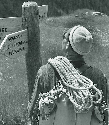 Une personne avec sac à dos et cordes regarde un panneau de randonnée en montagne.