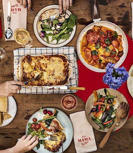 Une table bien garnie avec lasagnes, salades et pizza. Les mains atteignent la nourriture. Convivialité.