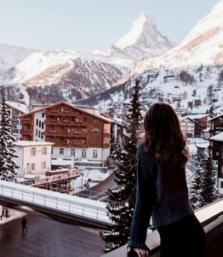 Eine Frau steht auf einem Balkon und blickt auf ein verschneites Bergdorf mit dem Matterhorn im Hintergrund.