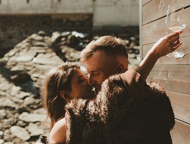 Couple in warm clothing embracing outside, woman holding a glass, snowy mountains in the background.