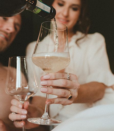 Man and woman enjoying a glass of sparkling wine on a bed.