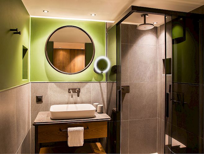 Salle de bain moderne en vert et gris, miroir rond, lavabo blanc et douche en verre.