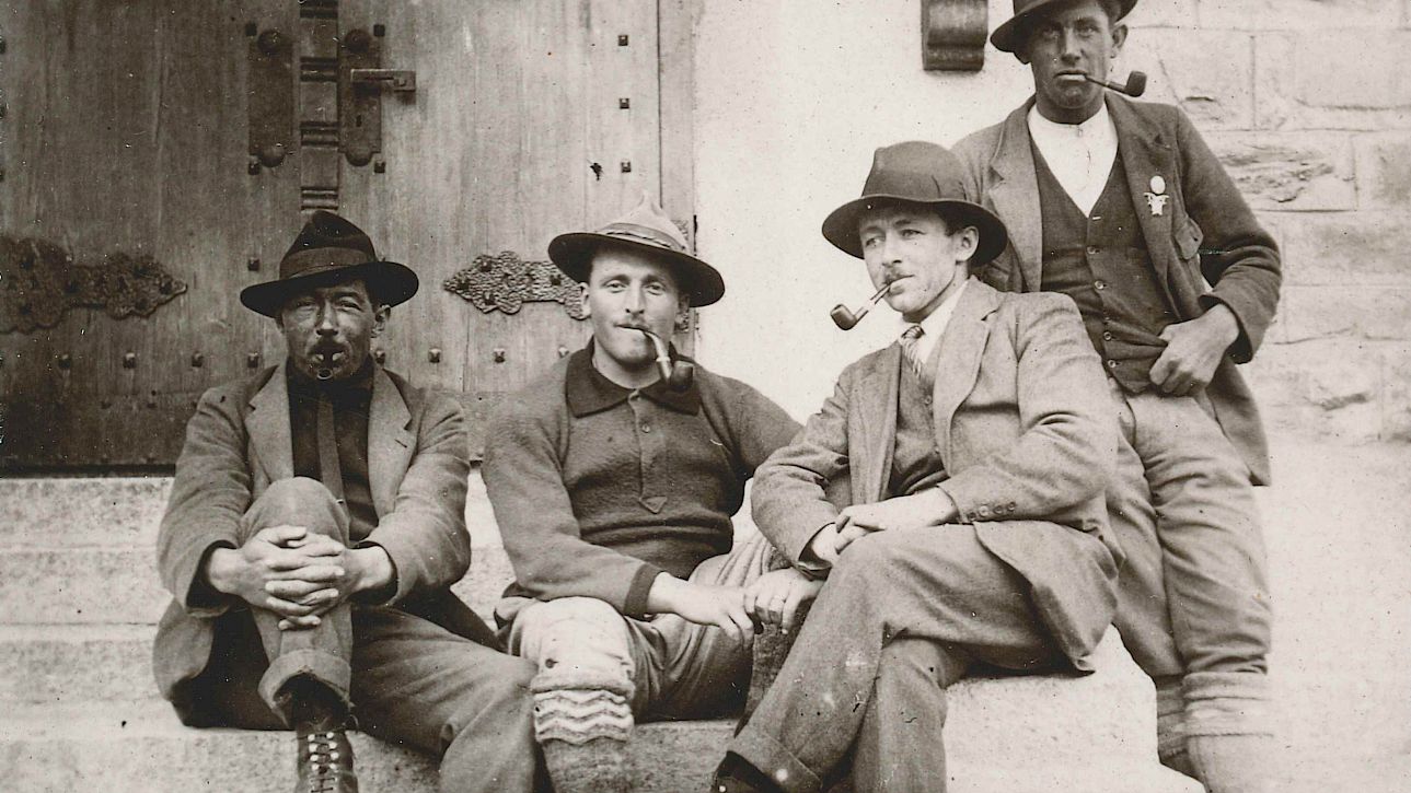 Quatre hommes en tenue traditionnelle assis sur des marches devant une porte en bois, fumant la pipe.