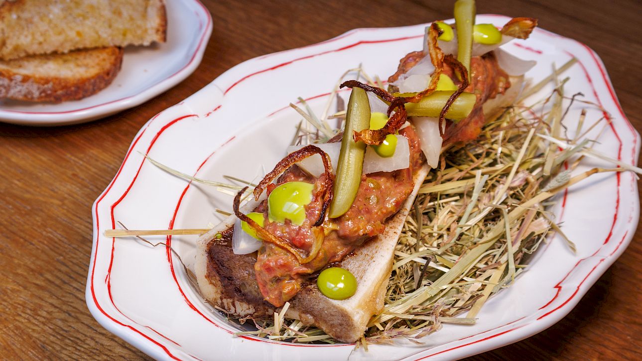 Creative appetizer with bread, pickles, and herbs on hay, served on a plate. Bread in the background.