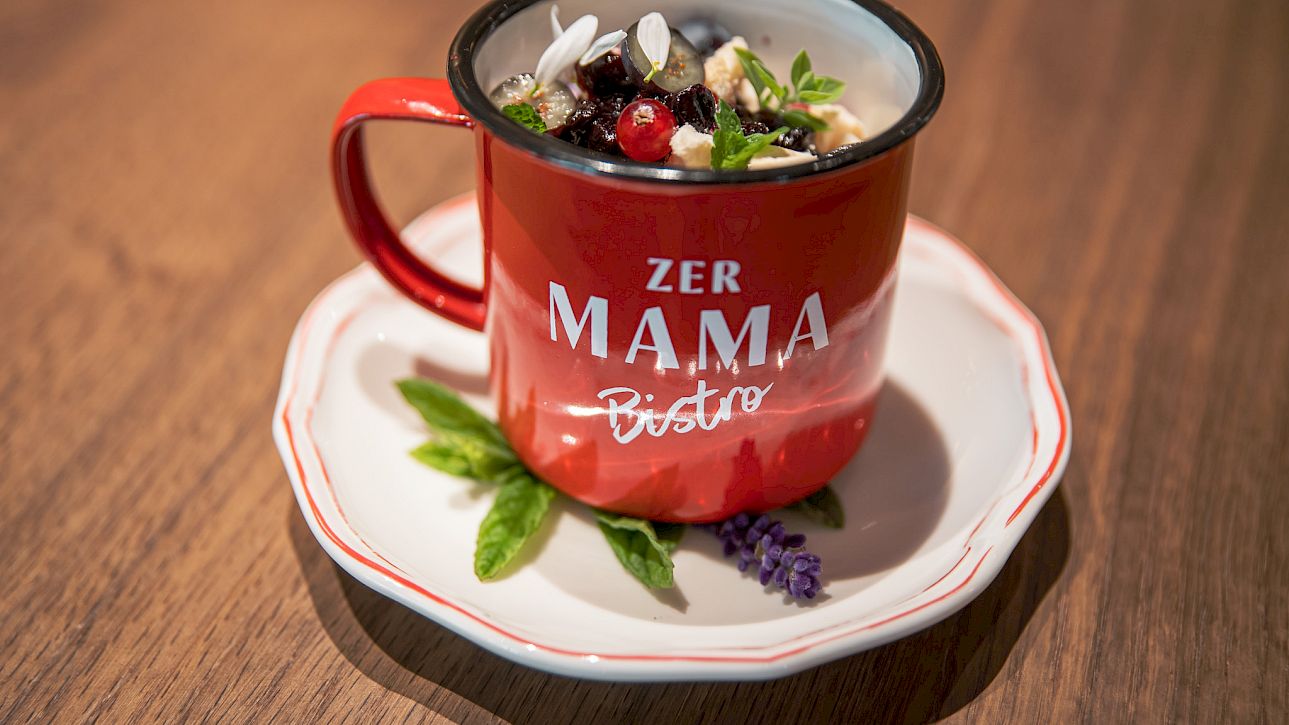 Red cup with "Zer Mama Bistro" text filled with yogurt and fruits on a decorative plate.