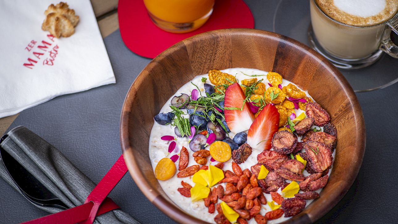 Wooden bowl with yogurt, fresh fruit, and nuts, next to a glass of juice and a cappuccino.