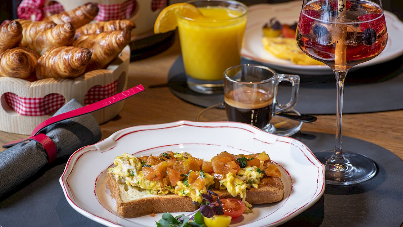 A rich breakfast table with croissants, toast with scrambled eggs, orange juice, and a glass of aperitif.
