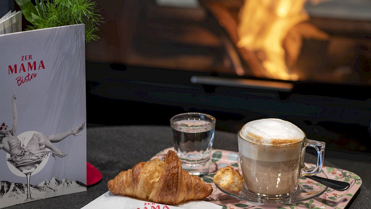 A croissant and cappuccino on a tray in front of a fireplace, "Zur Mama Bistro" menu in the background.