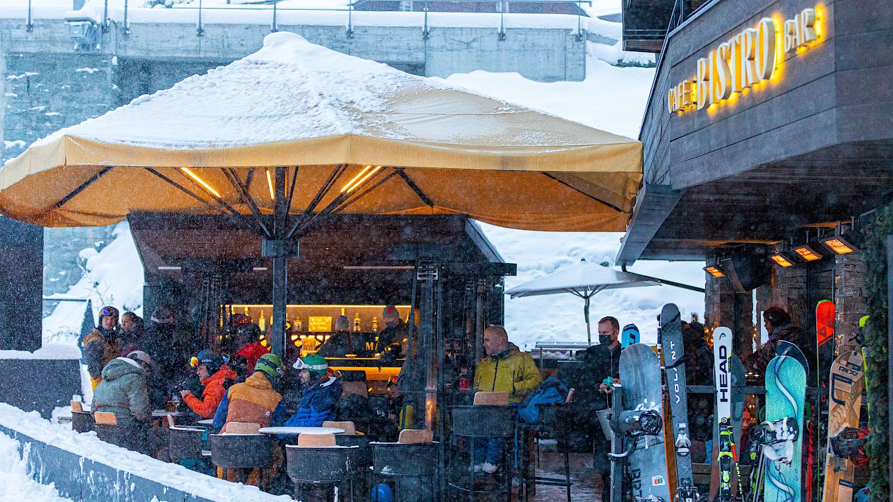 Ski outdoor bar in snow, people sitting under a yellow umbrella, with snowboards leaning nearby.