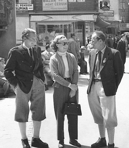 Three people smiling while talking on a street in front of shops. A woman and two men in traditional attire.