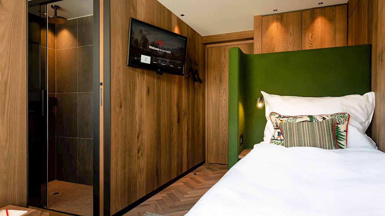 Modern hotel room with wooden walls, a bed with a green headboard, and a TV. Shower cabin in the background.