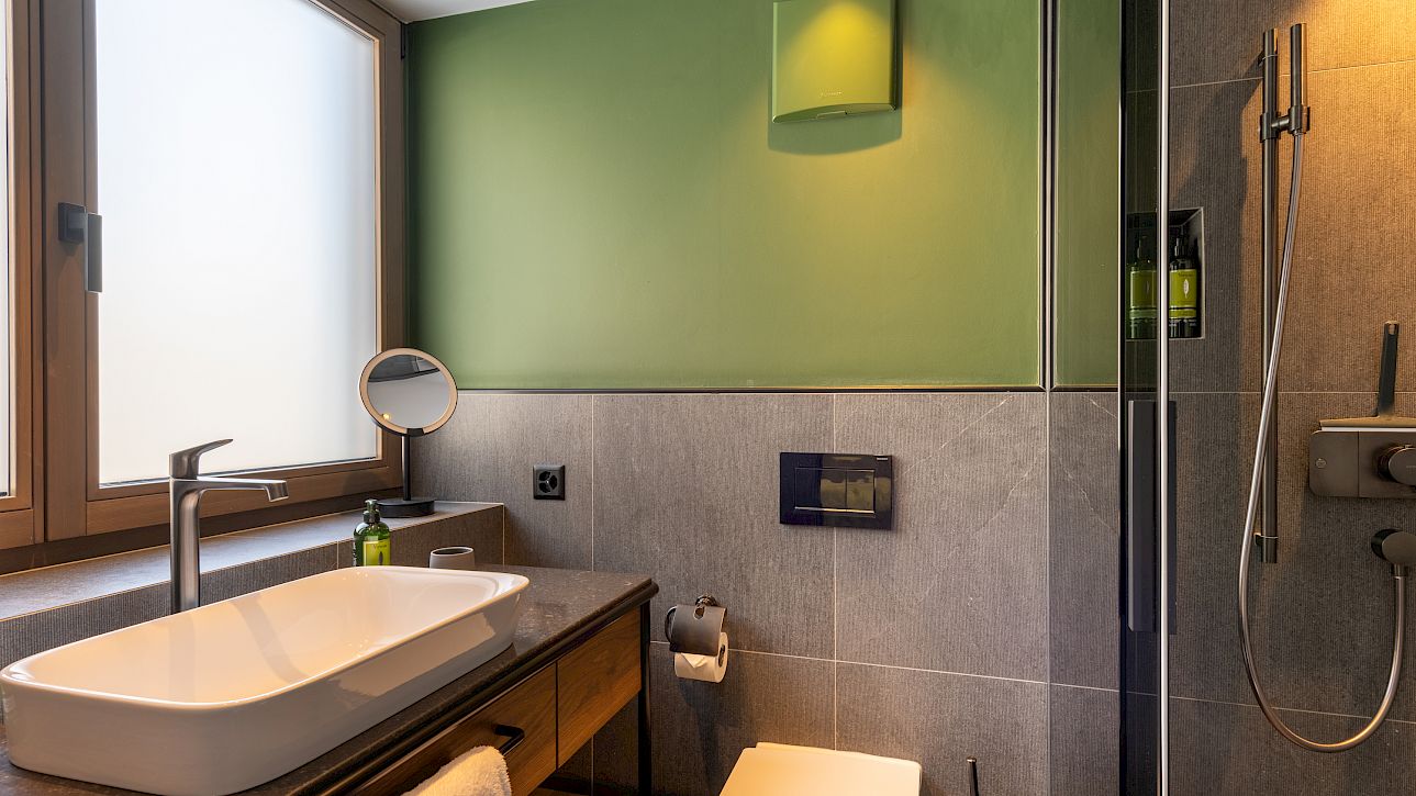 Modern bathroom with green wall, sink, mirror, and shower next to a large window.