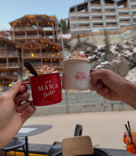 Two people clinking mugs with cream in front of alpine chalets; winter atmosphere.