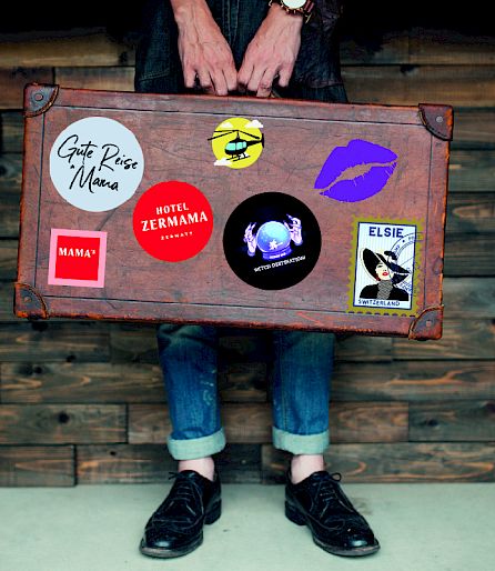 Person holds vintage suitcase with colorful stickers. Wooden wall background. Jeans and black shoes.
