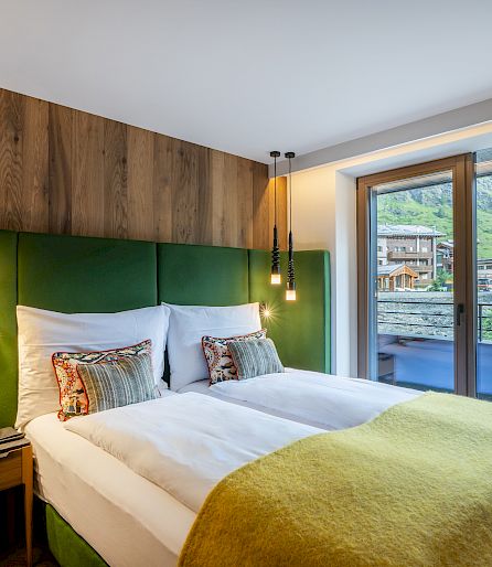 A modern hotel room with a large double bed, wood paneling, and a view of a mountainous landscape.