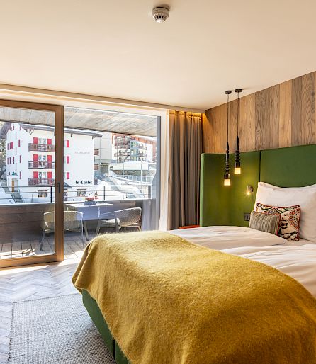 Cozy hotel room with double bed, wooden walls, and balcony overlooking snow-covered buildings.