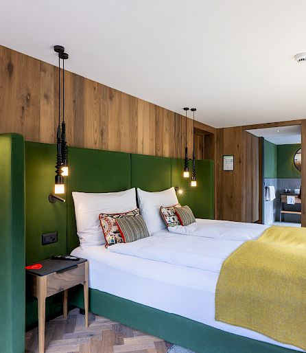 Cozy hotel room with double bed, wood paneling, and green accents, illuminated by daylight.