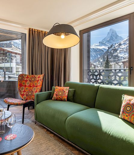 Living room with green couch, view of mountains and Matterhorn through large windows. Cozy atmosphere.