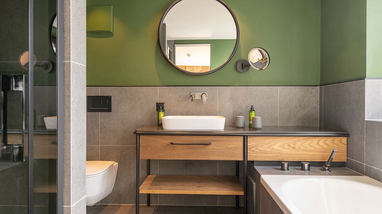 Modern bathroom with green walls, round mirror, wooden vanity, sink, and bathtub.