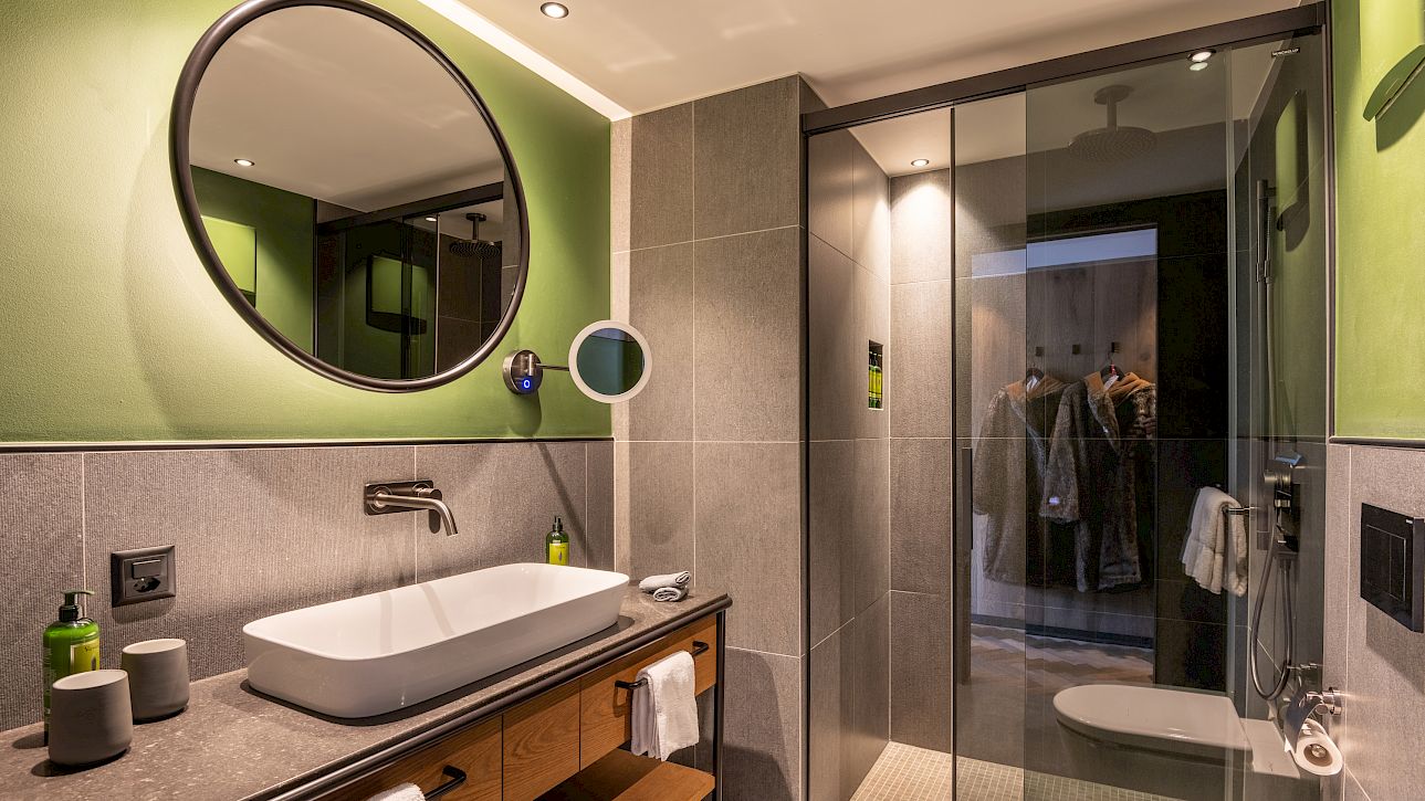 Modern bathroom with green-painted wall, round mirror, sink, and glass door to shower.