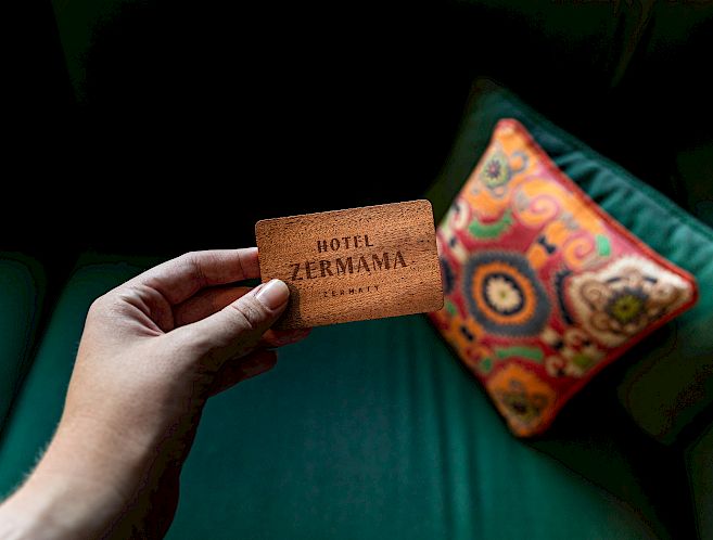 A hand holds a Hotel Zermama business card in front of a green-orange cushion on a sofa.