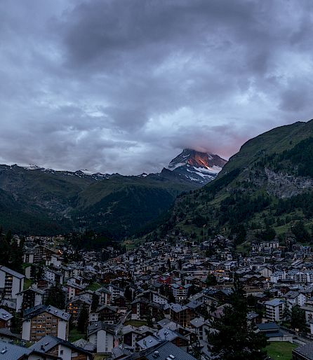 Cityscape at dusk, surrounded by mountains, with an illuminated peak in the background.