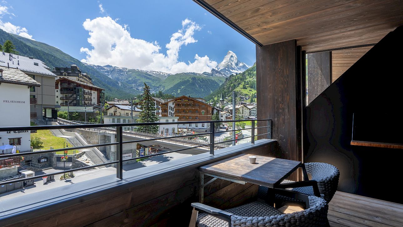 Balkon mit zwei Stühlen, Tisch und Bergblick auf sonnige Häuser in einem alpinen Dorf.