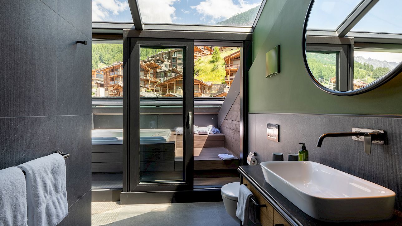 Modernes Badezimmer mit großem Fenster, das auf eine Terrasse zeigt, Außenwhirlpool und Bergblick.