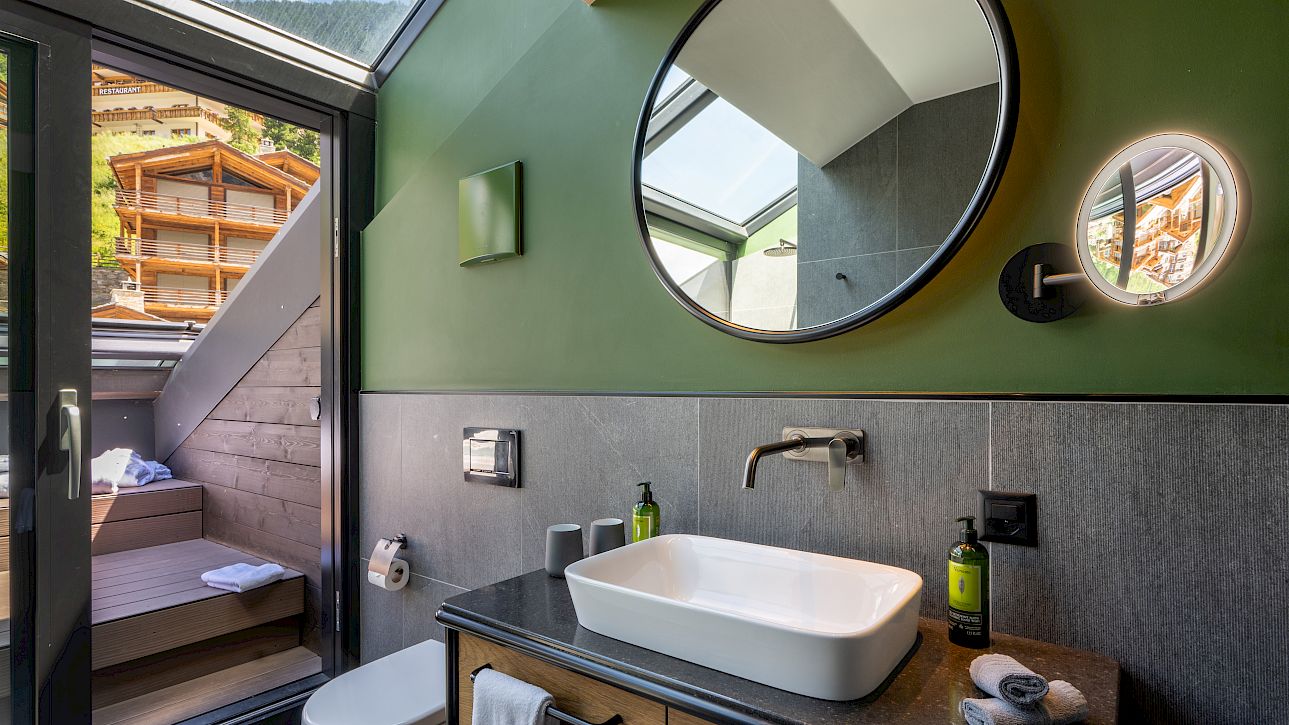 Modernes Badezimmer mit grünem Wandakzent, rundem Spiegel und Aussicht auf eine Terrasse mit Berghintergrund.