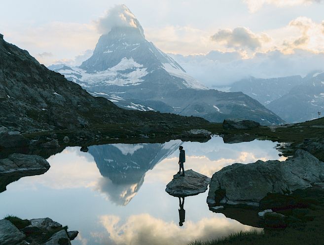 Berglandschaft mit See, in dem sich ein Berg spiegelt. Eine Person steht auf einem Felsen am Ufer.