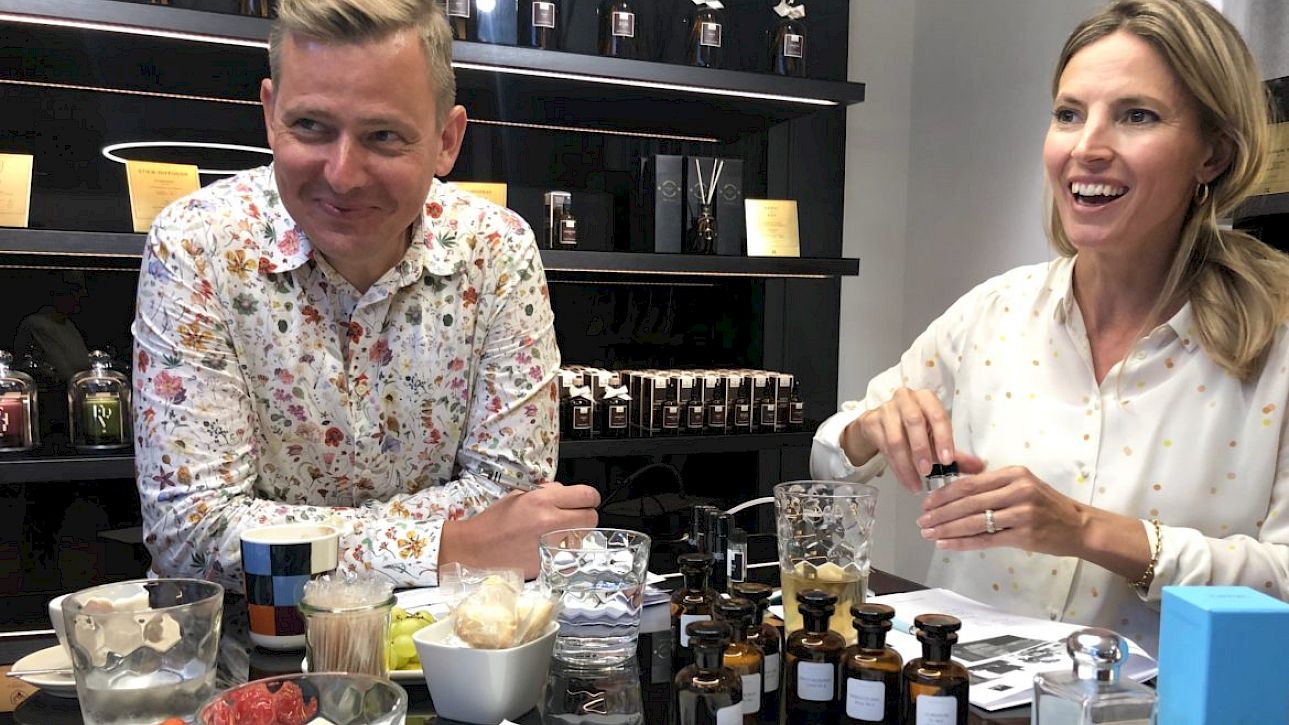Deux personnes souriantes, assises à une table avec des flacons de parfum et des ingrédients.