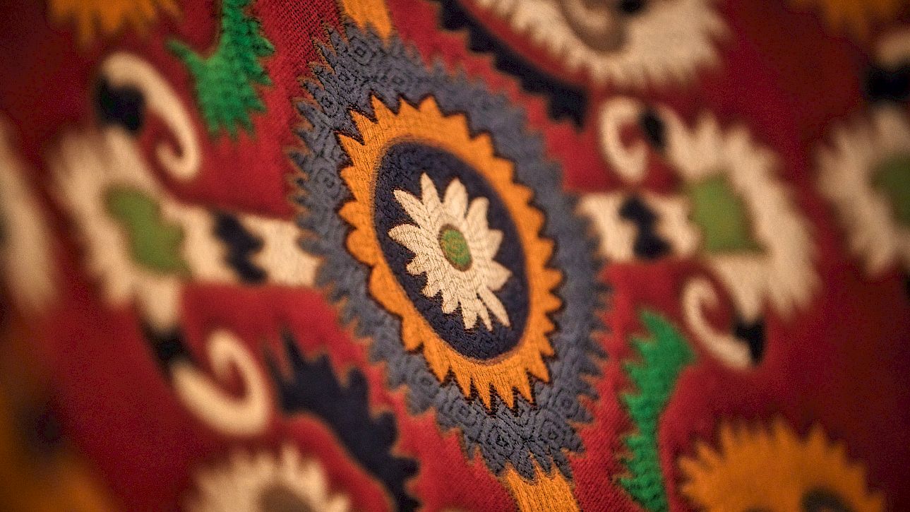 Tapisserie avec motifs colorés, incluant un motif floral central en bleu, jaune et blanc sur fond rouge.
