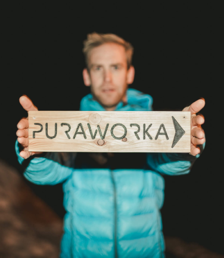 Ein Mann in blauer Jacke hält ein Holzschild mit der Aufschrift "PuraWorka" vor einem dunklen Hintergrund.