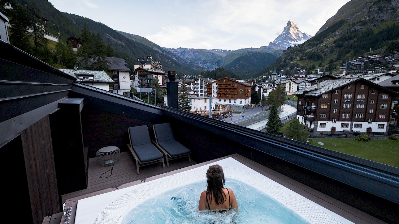Person relaxing in a rooftop hot tub overlooking a picturesque mountain town and snowy peaks.