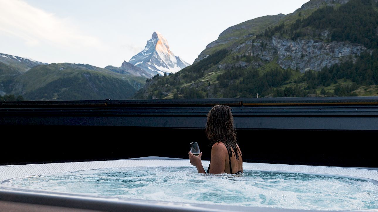 Person in hot tub gazing at mountains, holding a drink. Scenic view of snowy peak in the distance.