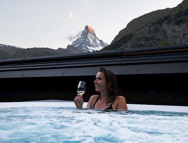 Woman enjoying a glass of wine in a hot tub with a mountain view at sunset.