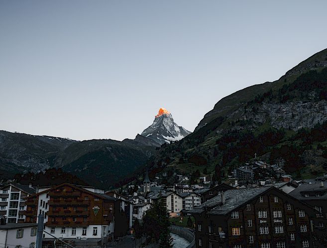 Cityscape at dusk with a lit mountain peak in the background, possibly the Matterhorn.