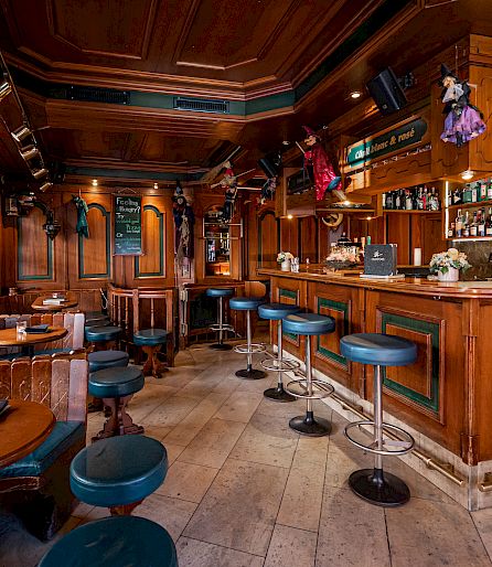 Cozy bar with wood-paneled walls, decorated shelves, and blue bar stools along the counter.