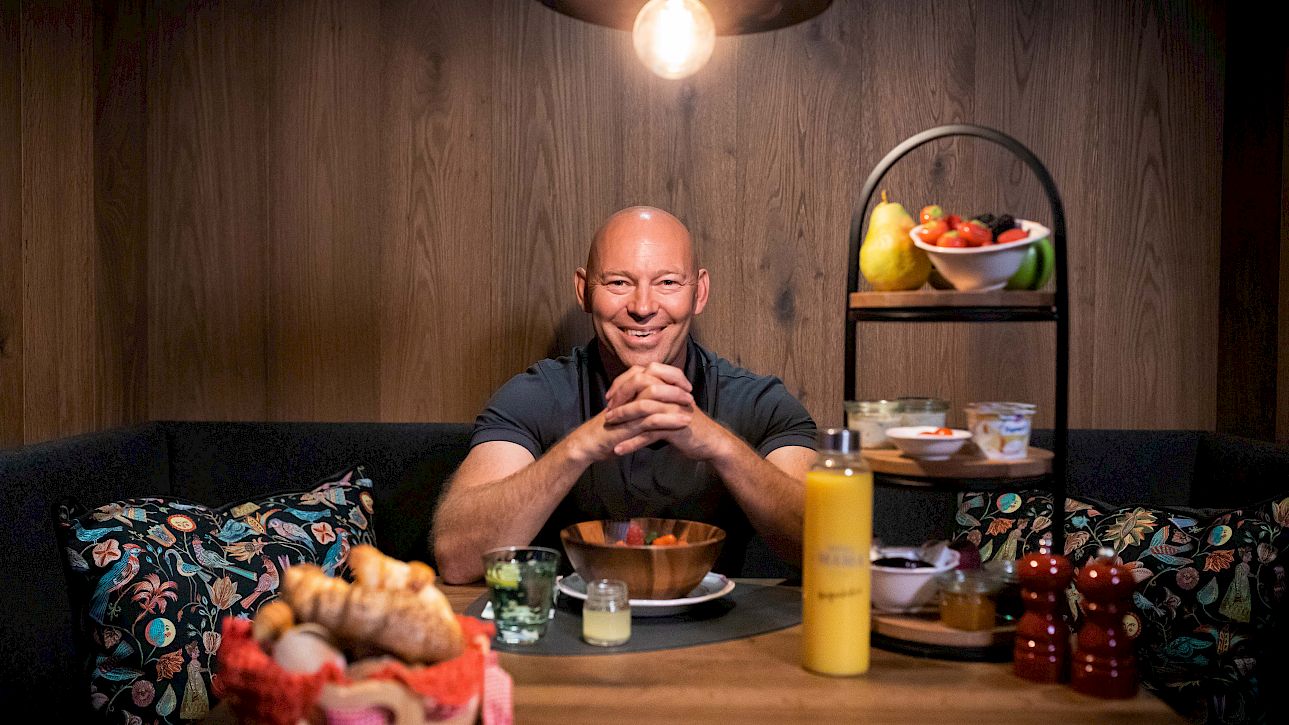 Un homme est assis à une table avec petit-déjeuner, y compris des petits pains, du jus et des fruits, dans une ambiance conviviale.