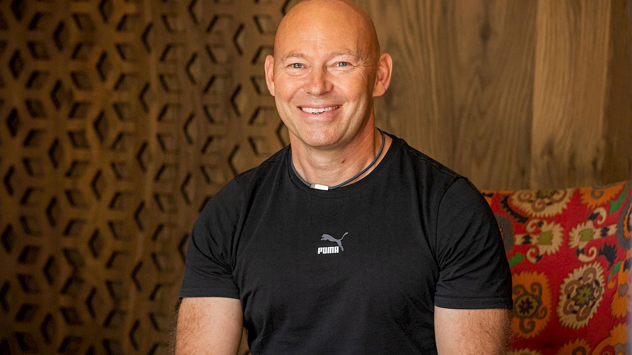 Un homme souriant en t-shirt noir est assis devant un mur en bois et un coussin coloré.