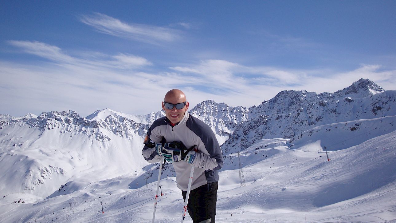 Un homme en tenue de ski se tient sur une piste enneigée avec des montagnes et un ciel bleu éclatant.