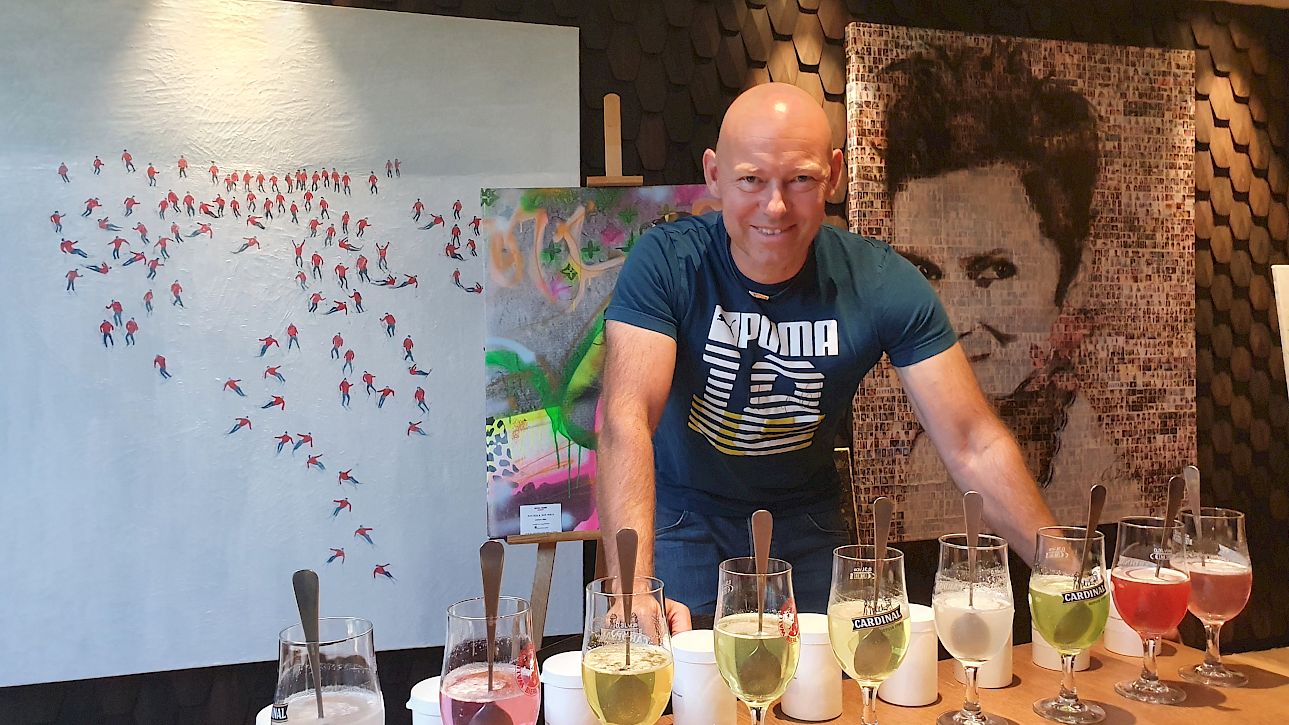 Un homme se tient derrière une rangée de verres avec des boissons colorées, œuvres d'art en arrière-plan.