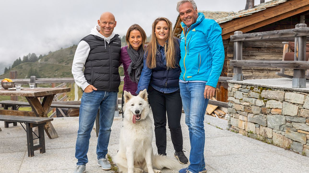 Quatre personnes et un chien blanc souriants posent devant un paysage de montagne près d'une cabane.