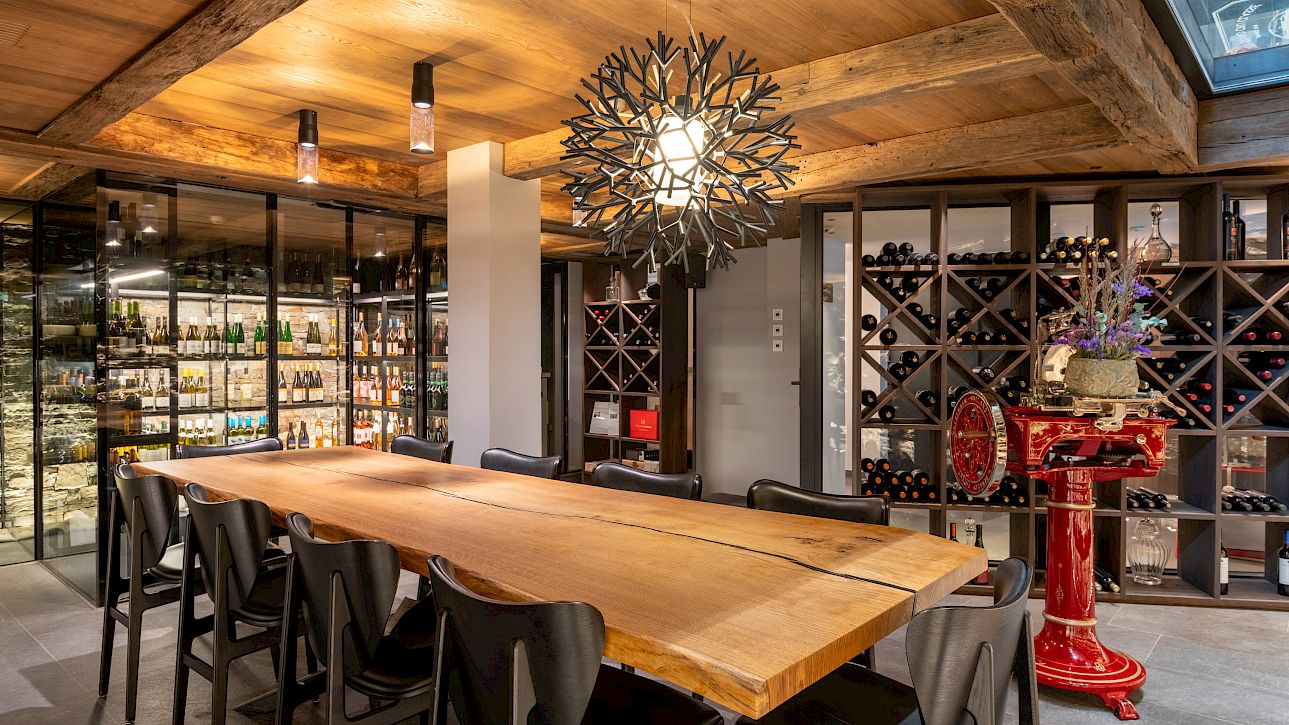 Cave à vin moderne avec grande table en bois, lustre design et étagères garnies de bouteilles de vin.