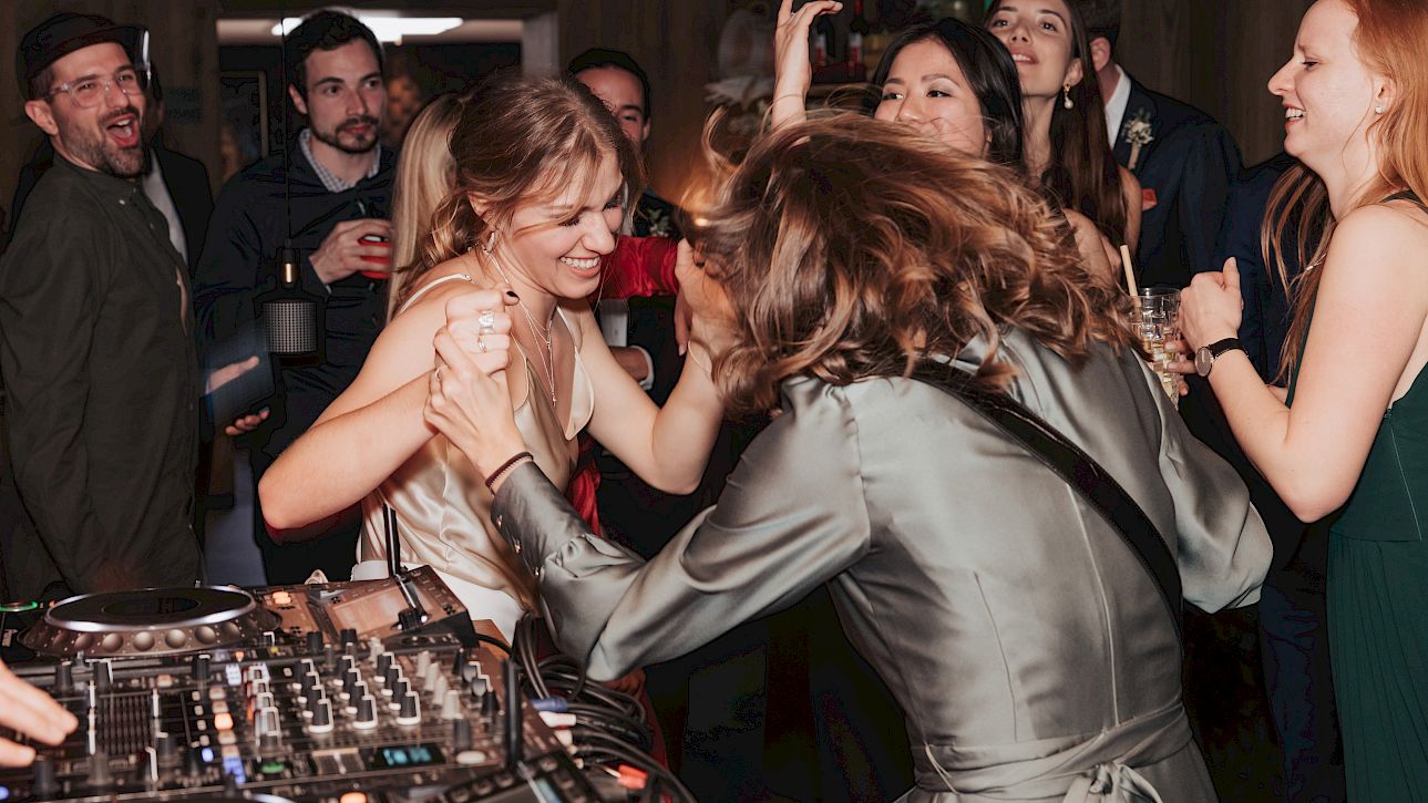 Deux femmes dansent joyeusement à une fête, entourées de personnes souriantes et d'une table de mixage au premier plan.