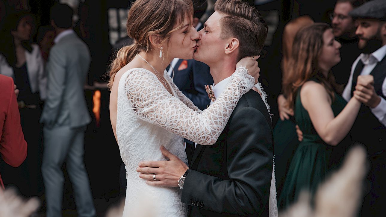 Les mariés échangent un baiser tendre lors de leur mariage, tandis que les invités dansent en arrière-plan.