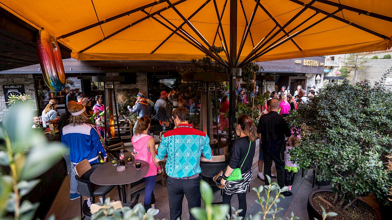 Des gens se rassemblent sous un grand parasol jaune sur une terrasse animée.