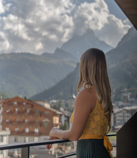 Frau in gelbem Oberteil betrachtet von einem Balkon aus die Berglandschaft.